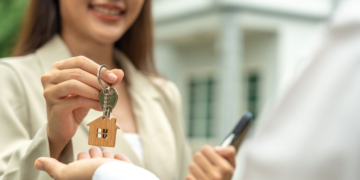 Smiling landlord handing over a house-shaped keychain to a tenant outdoors.