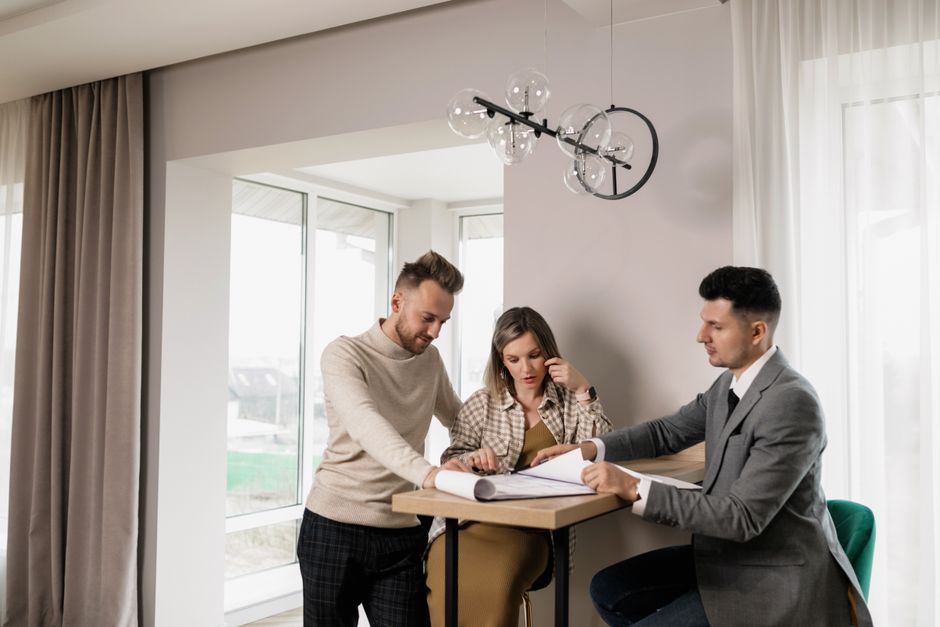 Smiling couple sits at their sunlit dining table, looking over papers related to their home sale, feeling confident and ha...